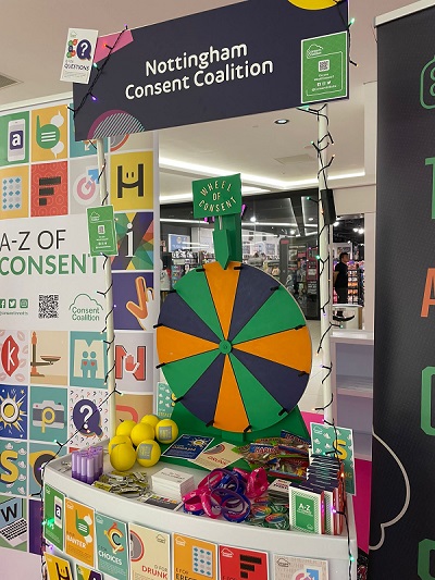 A photo of the Consent Coalition stall, featuring a 'wheel spinner game' prizes and a banner with the A-Z of Consent on it