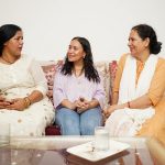 A photo showing three South Asian women on a settee. Two of them might be older sisters, while the other (sat in the middle) is a younger woman. The older woman on the left is talking, and the other are listening.