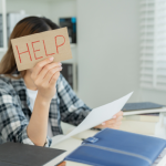 Woman sat at a desk, working, with on hand holding up a sign that says 'Help'