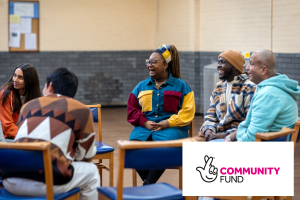 Picture of people of different genders and ethnicities sat in a circle laughing. In the bottom right is the National Lottery Community Fund logo, comprised of crossed hand image outlined in black and looking like a smiling face and 'Community Fund' written in pink and black.